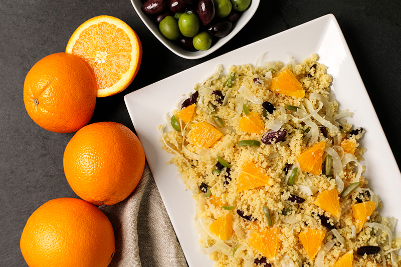 A platter of couscous arranged with oranges off to the side and a bowl of olives on top