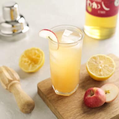 A glass of ume cocktail with halved citrus and a halved apple on a cuttingboard, ume wiskey off to the side