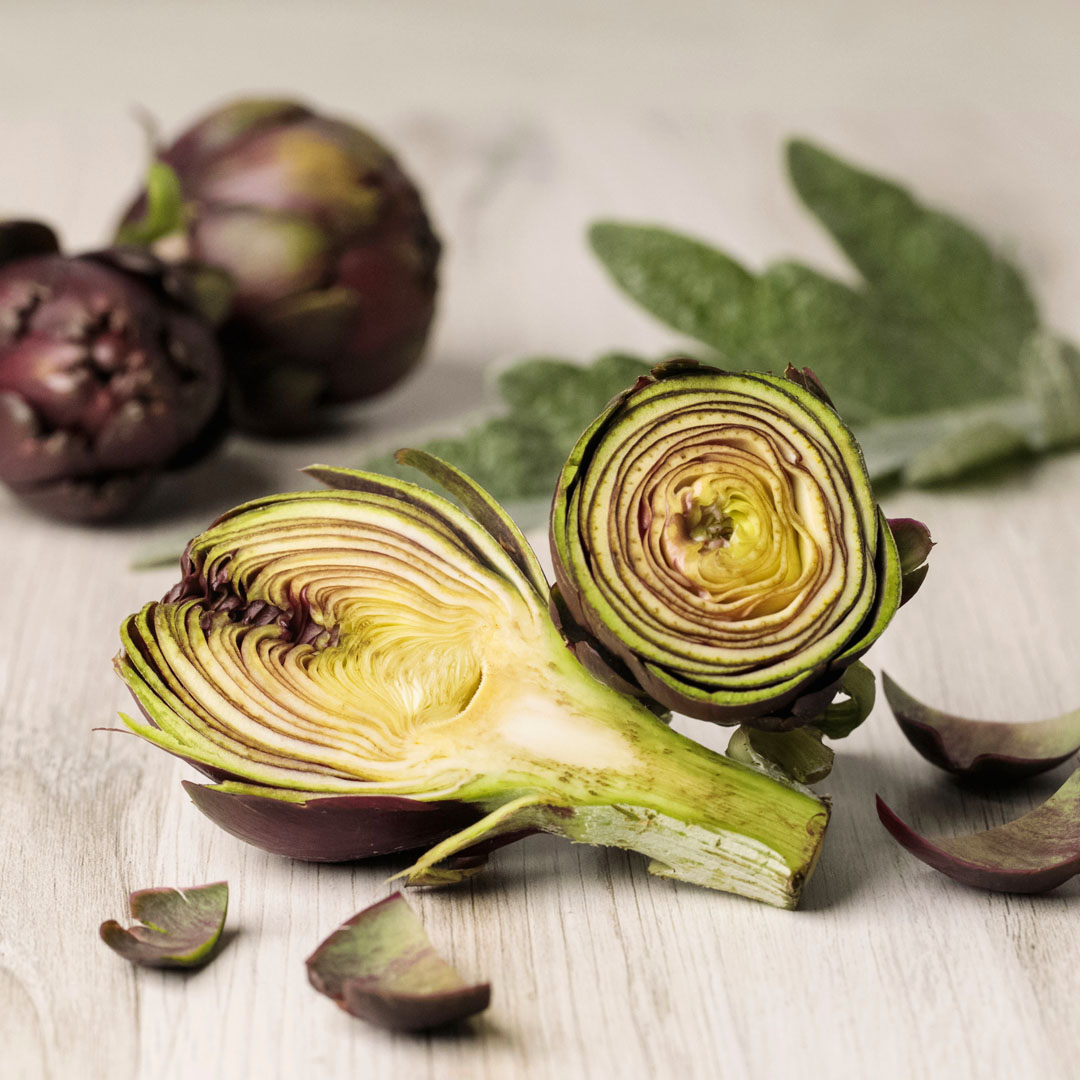An artichoke cut in halves, halves sitting on top of each other, whole flowers and leaves in background
