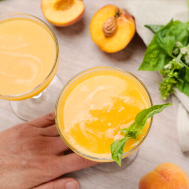 Hand holding coupe glass of blended peach mojito with surface covered with limes, peaches and basil leaves