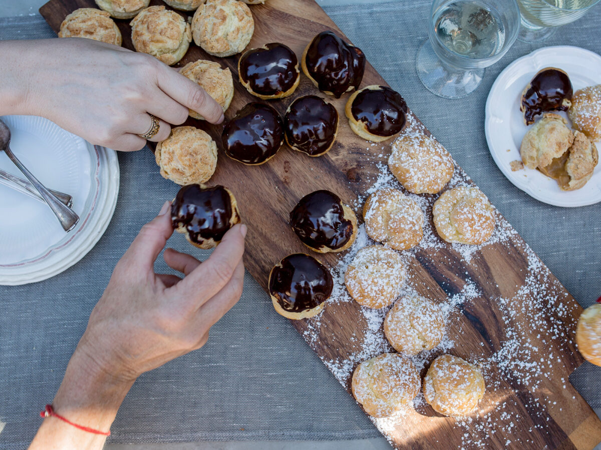 Two hands picking mini cream puffs off of a wooden serving board full of desserts