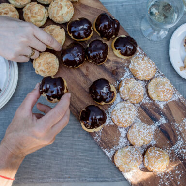 Two hands picking mini cream puffs off of a wooden serving board full of desserts