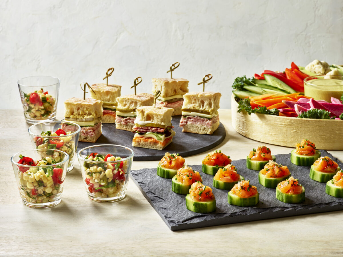 Various catered dishes, such as glass cups full of corn salad, a round slate of finger sandwiches, a slate of tuna tartare cucumber cups, a crudite platter in a wooden round