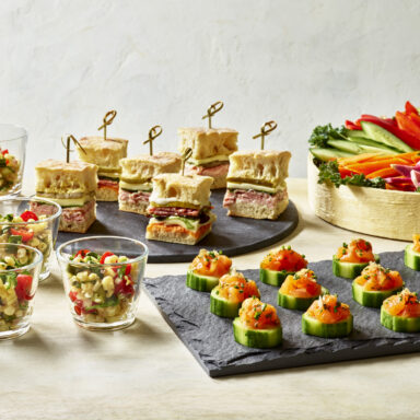 Various catered dishes, such as glass cups full of corn salad, a round slate of finger sandwiches, a slate of tuna tartare cucumber cups, a crudite platter in a wooden round
