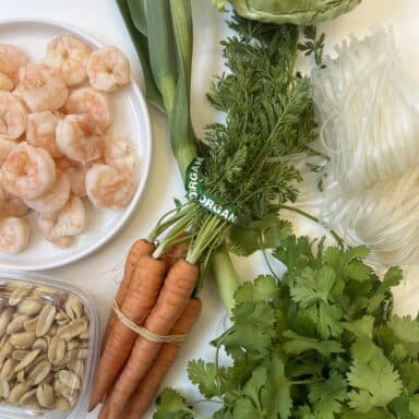 An overhead photo of grocery ingredients: noodles, carrots, cilantro, cabbage, shrimp, peanuts.