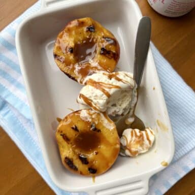 Two halves of grilled peaches in a white dish, topped with a scoop of ice cream and drizzled with caramel sauce.
