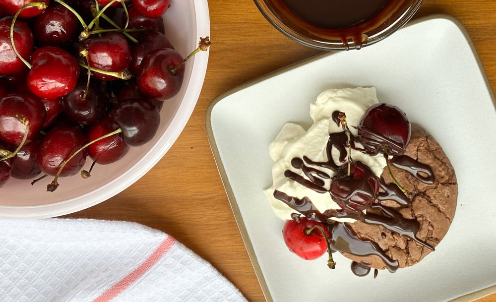 A chocolate cookie on a white plate, drizzled with chocolate sauce, with a dollop of whipped cream, and topped with fresh cherries.