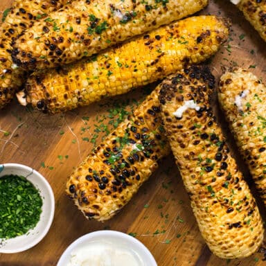 Eight ears of corn on a wooden surface, grilled and covered with herbs with herbs and cream on the side