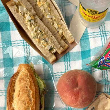 The egg salad sandwich that has been cut in half and there's a sandwich on a baguette with a peach, a bottle of sparkling water, and a bag of chips on a checkered tablecloth.
