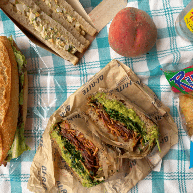 Three sandwiches, a peach, a bottle of sparkling water, and a bag of chips on a checkered tablecloth. r
