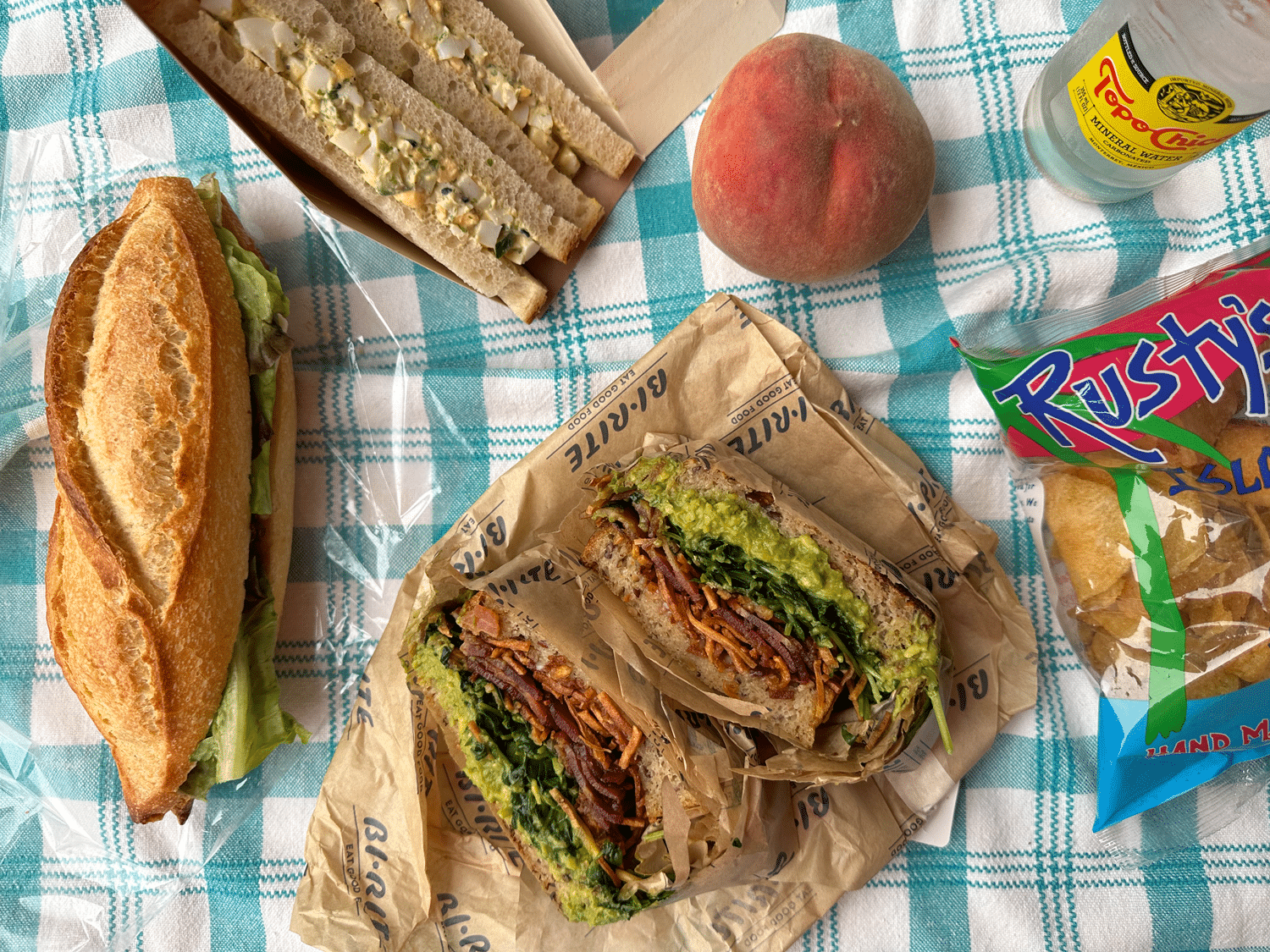 Three sandwiches, a peach, a bottle of sparkling water, and a bag of chips on a checkered tablecloth. r