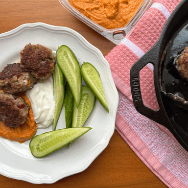 Garlicky meatballs served on a white plate with a dollop of hummus and yogurt with a side of cucumbers.