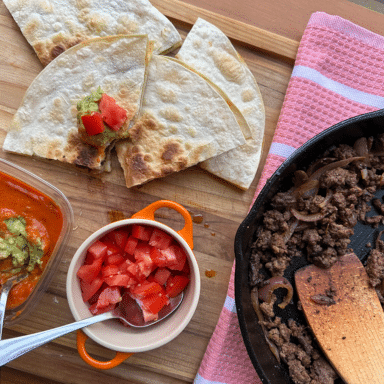 Quesadillas being served with tomatoes and guacamole on a wooden cutting board, next to a cast iron skillet with ground beef.