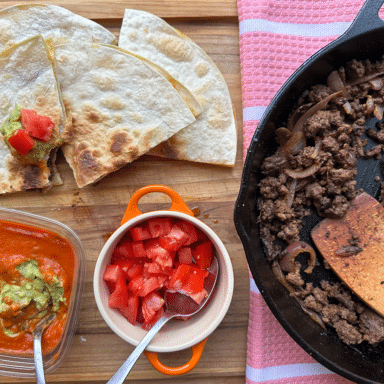 Quesadillas being served with tomatoes and guacamole on a wooden cutting board, next to a cast iron skillet with ground beef.