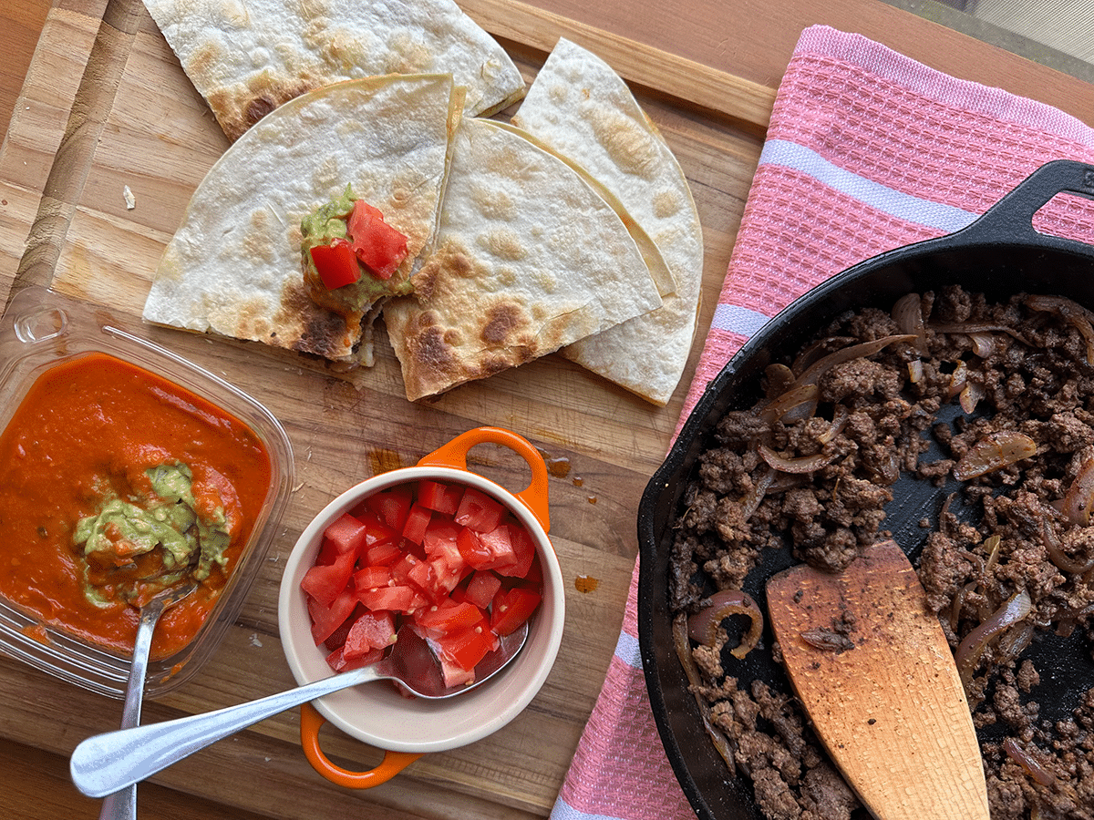 Quesadillas being served with tomatoes and guacamole on a wooden cutting board, next to a cast iron skillet with ground beef.