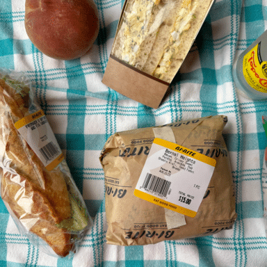 Three sandwiches in wrappers, a peach, a bottle of sparkling water, and a bag of chips on a checkered tablecloth.