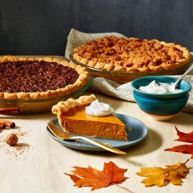 A whole pecan pie and apple pie, and a slice of pumpkin pie pulled out on a plate, with a dollop of whipped cream on top.