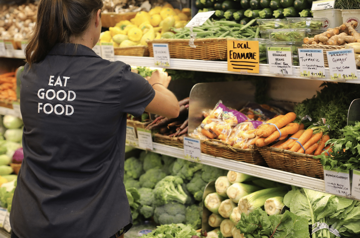 A staff member restocking a produce case, which is filled with carrots, celery, and broccoli.