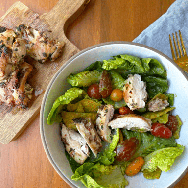 Herbed chicken thighs on a wooden cutting board, next to a bowl of salad with sliced chicken on top.