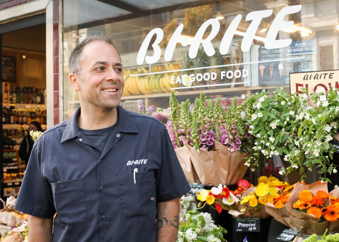 Sam standing and smiling in front of the Bi-Rite storefront, backed by bouquets of flowers.