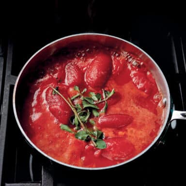 A Saucepan Filled With Whole Peeled Tomatoes And A Sprig Of Herbs, Coming Up To A Simmer On The Stove
