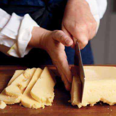 Hands Slicing A Slab Of Thick, Cooled Polenta With A Cleaver On A Cutting Board