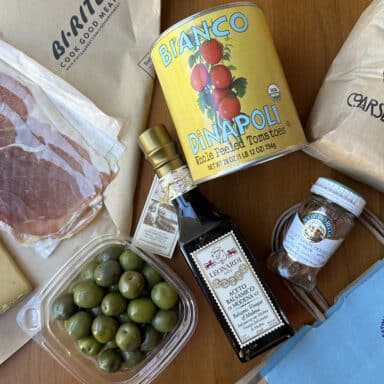Unpacking a Bi-Rite grocery bag, with packages of prosciutto, tomatoes, polenta, cheese, olives, vinegar, and anchovies spilling out on a table.