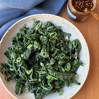 A Shallow Bowl Filled With Sautéed Kale Leaves, With Spoon Digging Into A Jar Of Bright Red Chili Crisp
