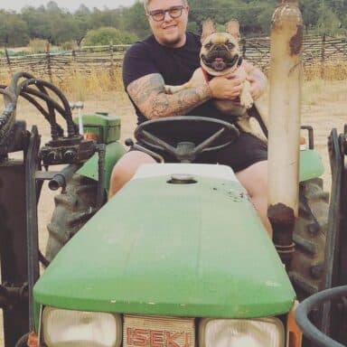 A Farmer And A Tan Bulldog Riding On A Green Tractor Together And Smiling