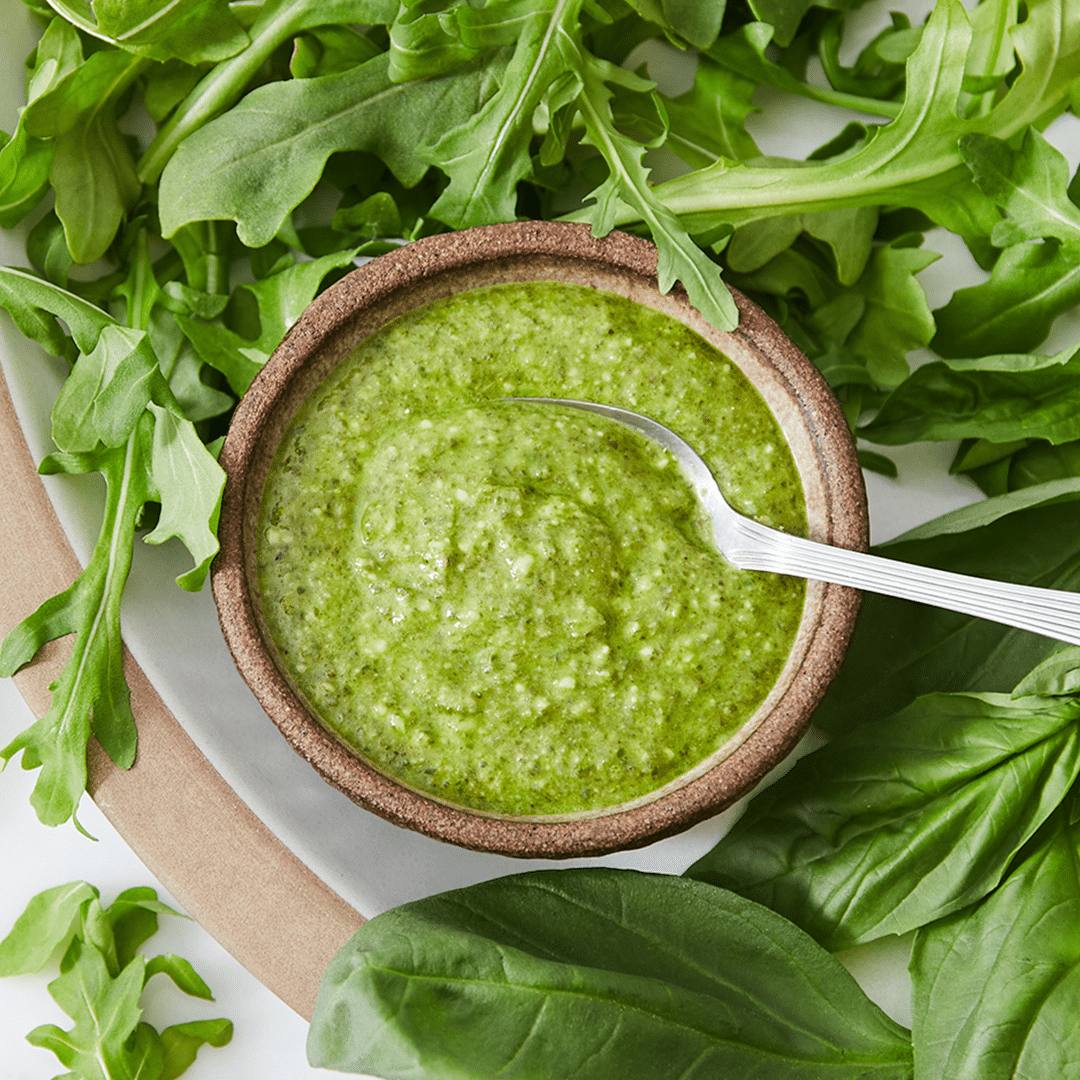 Any Green Pesto in a bowl shot top down surrounded by basil and arugula