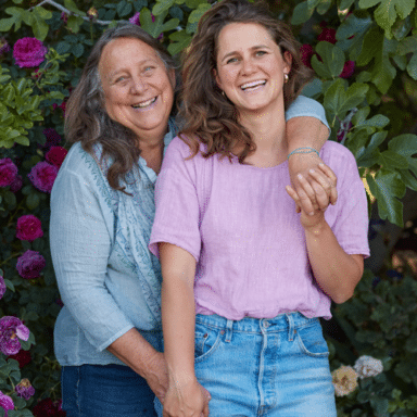 Laughing Dru Rivers And Hannah Muller Of Full Belly Farm, Facing The Camera, Dru's Arm Draped Across Hannah Amongst A Lush Canopy Of Flowers