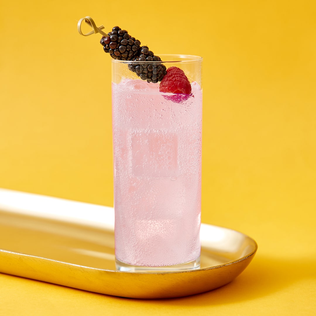 A gin cocktail in a highball glass with a berry garnish on a yellow background