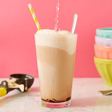 Ice cream float in a glass with soda pouring into it, a stack of four pastel bowls on the right, ice cream scoop on the left