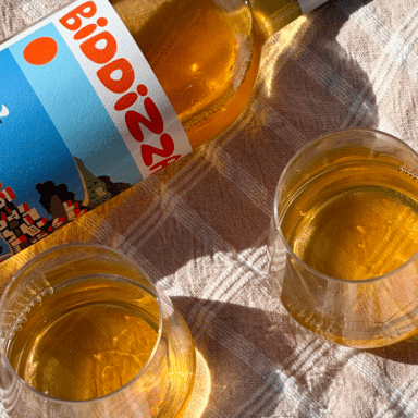 A Bottle Of Biddizza Laid Down On A Picnic Blanket Next To Two Full Glasses