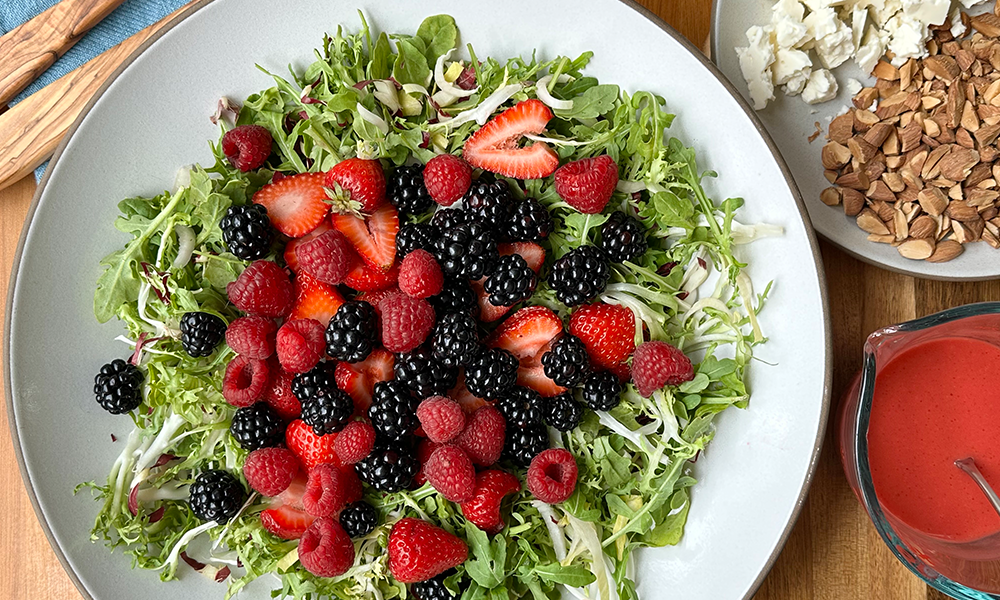 Summer Berry Salad Plated With Frisee And Chese And Almonds And Dressing On The Right Side With Blue Napkin And Tongs